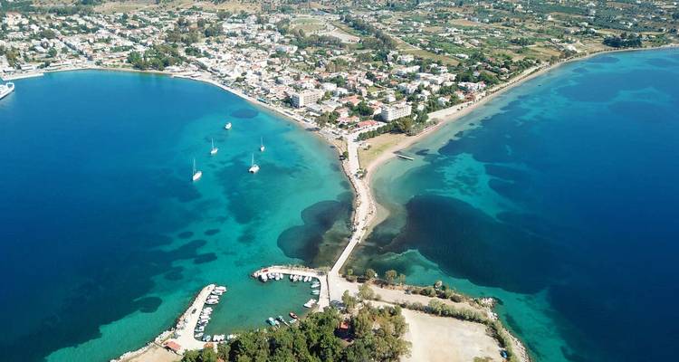 Vista aérea de una calzada estrecha que conecta dos mitades de un pueblo costero rodeado de bahías de azul vívido y veleros amarrados.