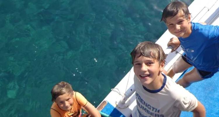 Drei nasse Kinder lächeln von der Seite eines Bootes auf, das über klarem, türkisblauem Wasser vor Anker liegt.