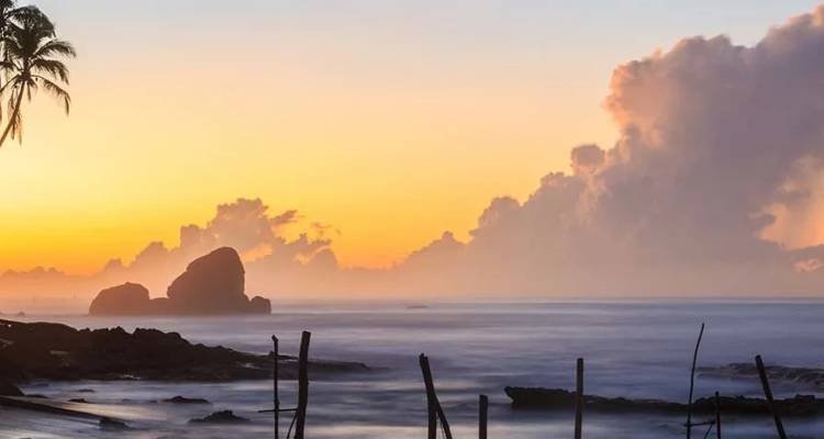 Playa tropical al amanecer con cielo anaranjado, olas suaves y rocas perfiladas