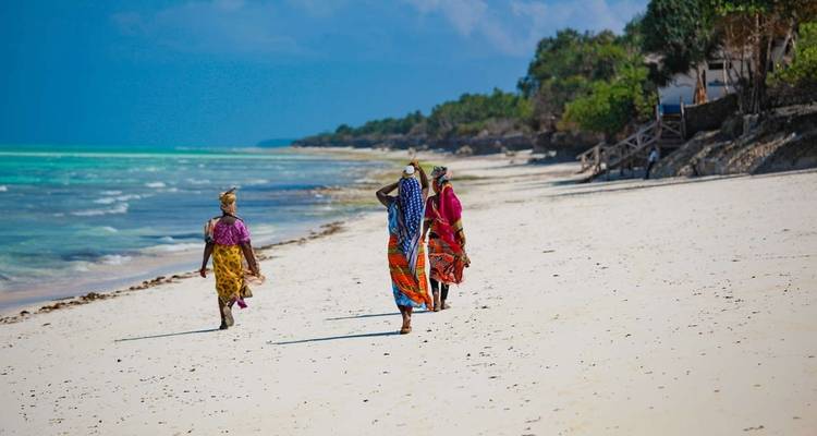 Drei Frauen in bunten Kleidern gehen an einem sandigen Strand entlang.