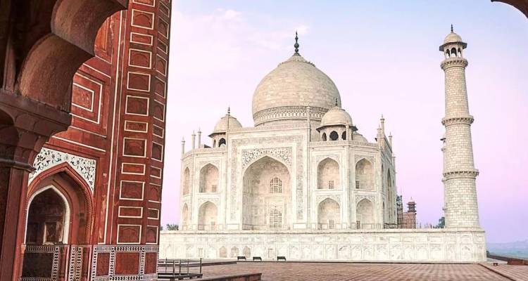 El icónico Taj Mahal de mármol blanco enmarcado por un arco de arenisca roja al amanecer con un cielo rosa suave.