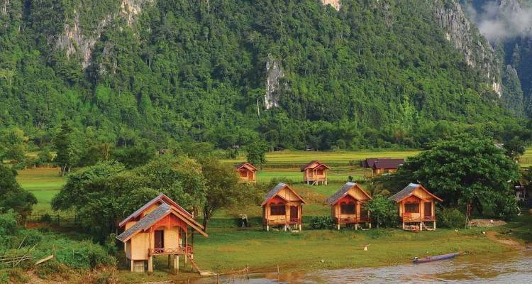 Rangée de petites cabanes en bois au bord de la rivière, dominées par d'imposantes falaises de calcaire vert et une jungle dense à Vang Vieng.