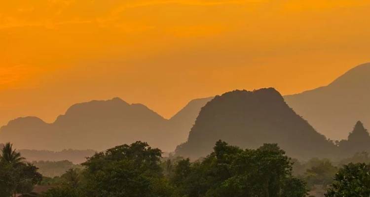 Las colinas de piedra caliza recortadas contra el cielo se elevan sobre la selva brumosa y los árboles en un cálido cielo de puesta de sol de color naranja de Laos.