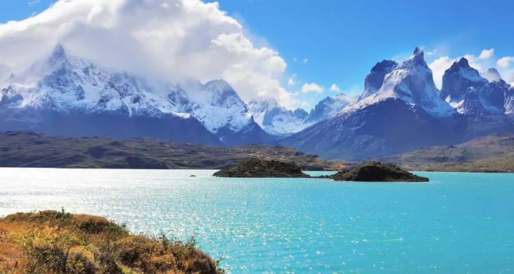 Sneeuwbedekte granieten torenspitsen torenen uit boven een turquoise meer en glooiende Patagonische heuvels onder dramatische wolken.