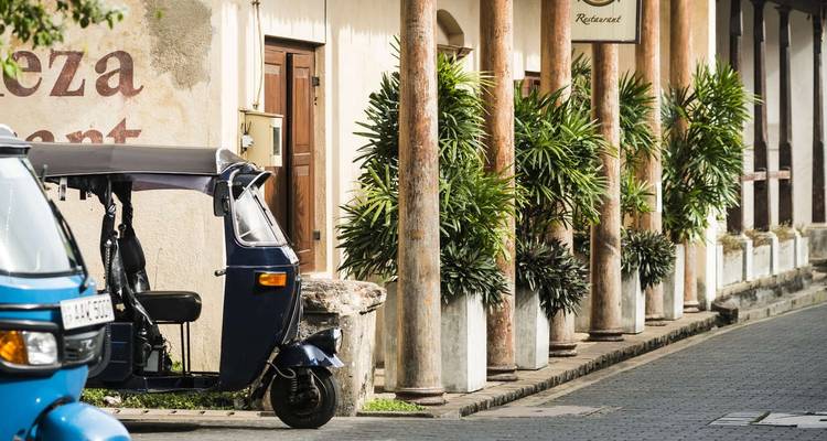 Un tuk-tuk bleu garé à côté de colonnes de style colonial et de palmiers en pots le long d'une rue tranquille.