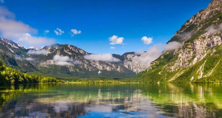 Un lago alpino cristalino que refleja bosques verdes y dramáticas montañas eslovenas bajo un cielo azul vivido con nubes dispersas.