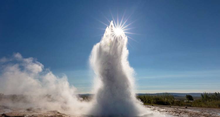 Un geyser puissant en éruption avec une explosion de soleil à son sommet sur un ciel bleu clair islandais.