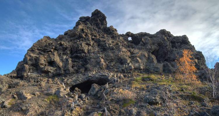 Formations de lave accidentées et entrée de grotte à Dimmuborgir sous un ciel bleu parsemé.