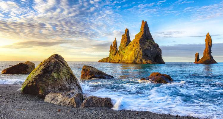 Les cheminées marines de Reynisdrangar au large d'une plage de sable noir illuminée par la lumière chaude du lever de soleil avec des vagues déferlantes au premier plan.
