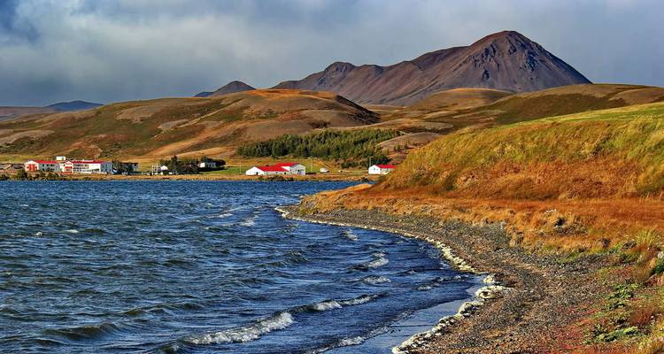 L'eau ondulante du lac Myvatn avec un petit village et des bâtiments aux toits rouges situés sur fond de collines onduleuses.