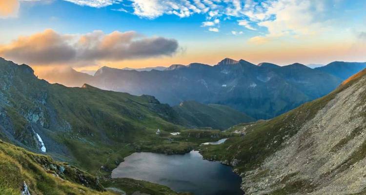 Panorama de gran altitud de un lago alpino rodeado de empinadas laderas verdes y picos rocosos al amanecer.