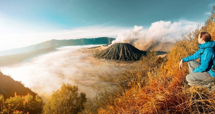 A person sitting on a hill overlooking an active volcano spewing smoke.