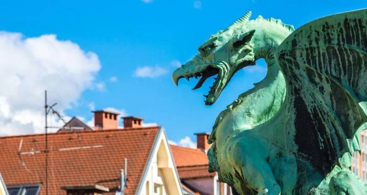 Weathered green dragon statue snarls above the rooftops of Ljubljana’s Old Town against a bright blue sky.