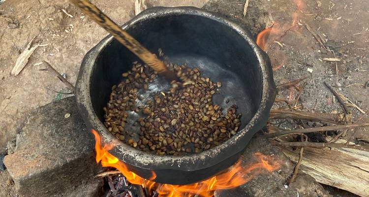 Eine Hand rührt Kaffeebohnen um, die in einem schwarzen Topf über einem offenen Holzfeuer geröstet werden.