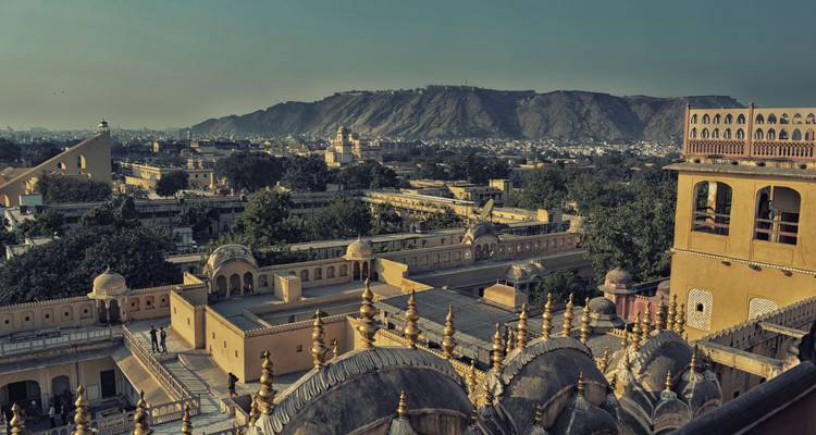 Una vista elevada de los tejados y cúpulas ornamentados de Jaipur con las colinas Aravalli formando un fondo brumoso bajo la cálida luz del atardecer.