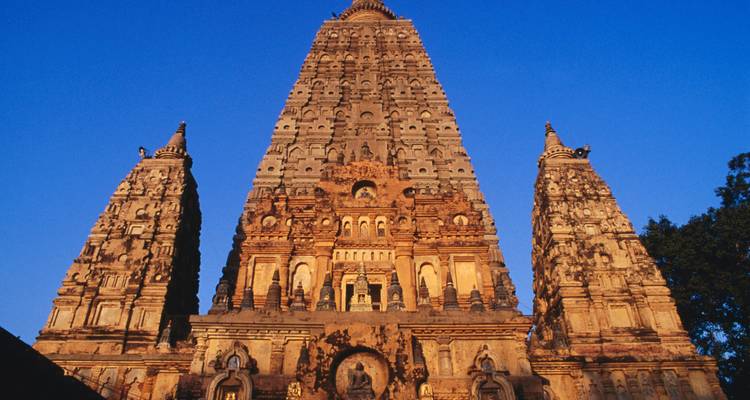 Una vista de ángulo bajo de la aguja principal del Templo de Mahabodhi y sus torres laterales brillando cálidamente contra un cielo azul profundo.