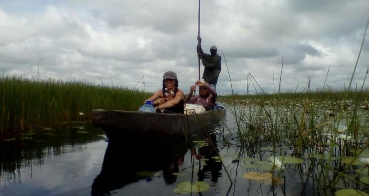Dos personas en una canoa navegando por agua con juncos y cielo nublado.