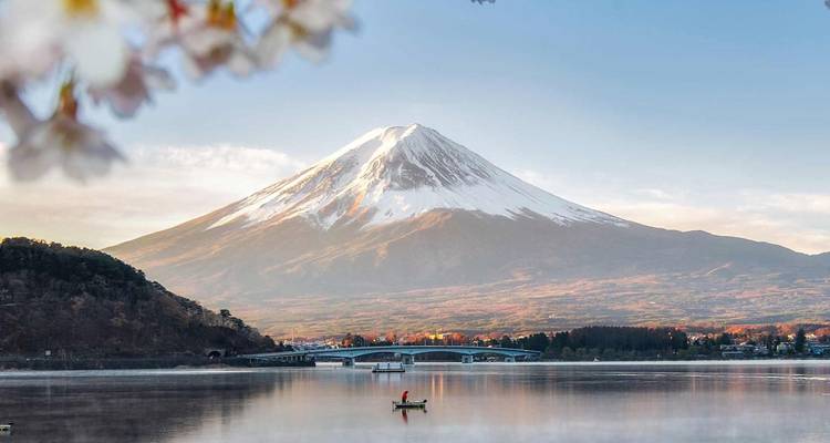 El Monte Fuji con cumbres nevadas se refleja en un lago tranquilo enmarcado por flores de cerezo al amanecer.