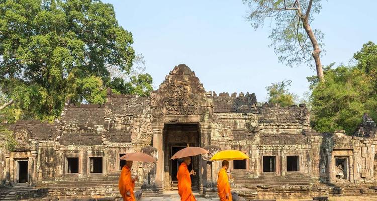 Tres monjes vestidos con túnicas naranjas y paraguas caminan junto a la entrada de un antiguo templo de piedra.