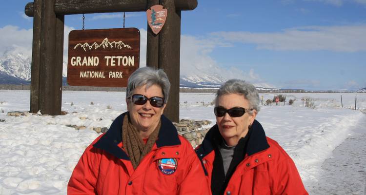 Deux femmes souriantes en vestes rouges se tiennent devant le panneau d'entrée du parc national de Grand Teton.