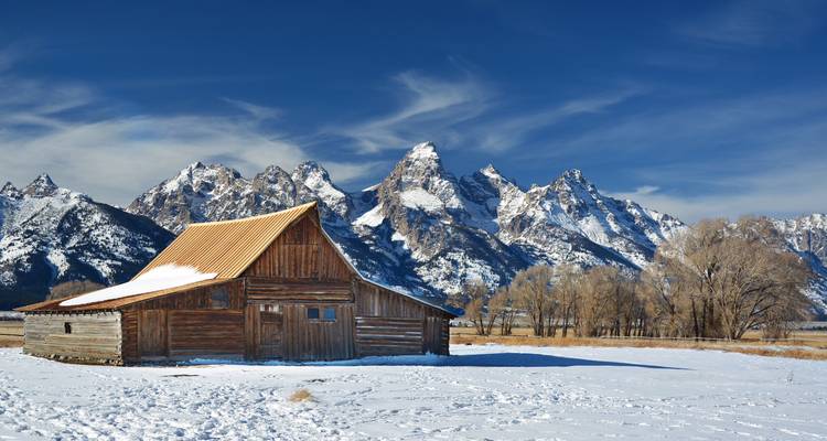 Une grange historique en bois se dresse devant la chaîne dentelée des Tetons aux sommets enneigés sous un ciel bleu éclatant.