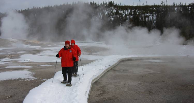 Deux personnes en parkas rouges font de la raquette en passant devant des évents géothermiques fumants sur une promenade en bois hivernale.