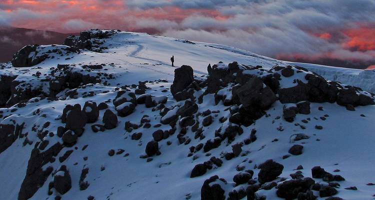 Person, die bei Sonnenuntergang auf einem verschneiten Berggrat wandelt.