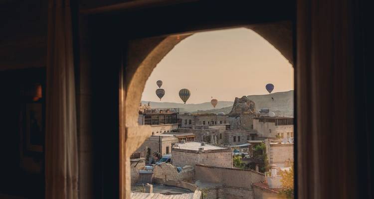 Los globos aerostáticos flotan sobre casas de piedra y chimeneas de hadas, vistos a través de una ventana arqueada al amanecer.