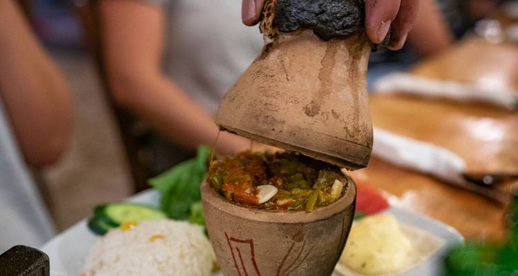 Una mano abre una olla de barro revelando un guiso turco humeante junto a arroz y ensalada.
