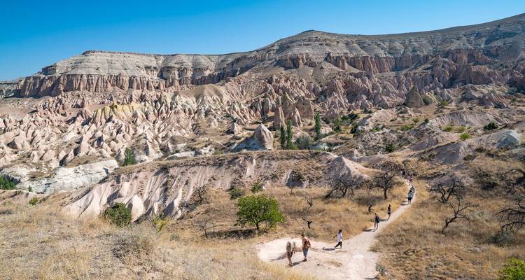 Los caminantes siguen un sendero polvoriento a través de los escarpados valles de roca rosada de Capadocia bajo cielos despejados.
