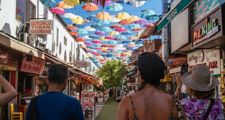 Los turistas pasean por una calle animada techada con coloridos paraguas colgantes entre tiendas y cafés.