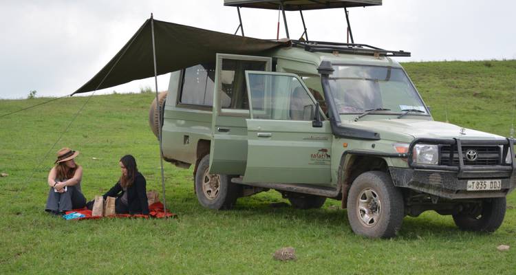 Two women sitting under a canopy next to a safari vehicle.