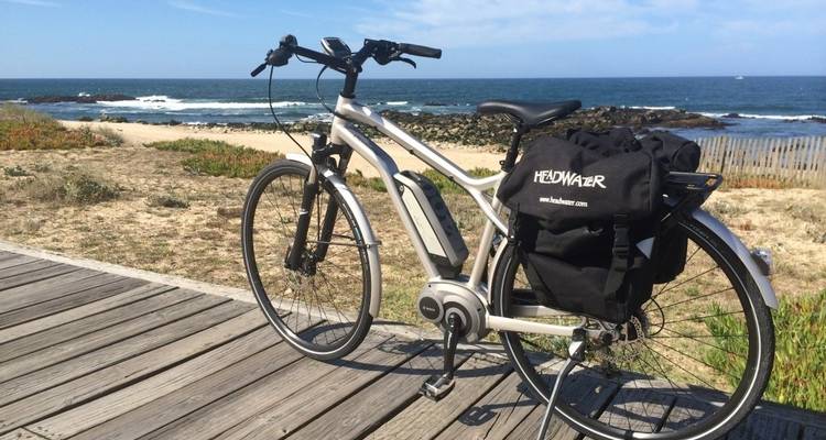 Una bicicleta de turismo descansa sobre un paseo marítimo de madera con vista a una playa arenosa del Atlántico y olas ondulantes.