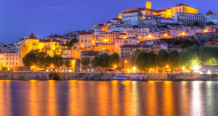 Los edificios ribereños de Coimbra iluminados coloridamente se reflejan en las aguas tranquilas del atardecer bajo un cielo crepuscular violeta.