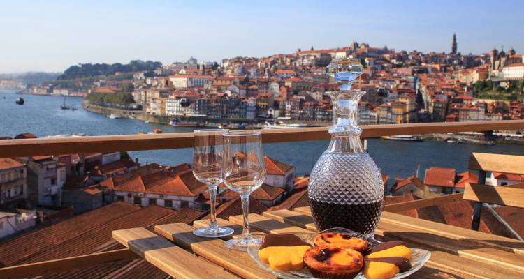 Una garrafa de vino de Oporto, dos copas y pasteles locales reposan en una terraza con vista al horizonte histórico de Oporto y al río Duero.