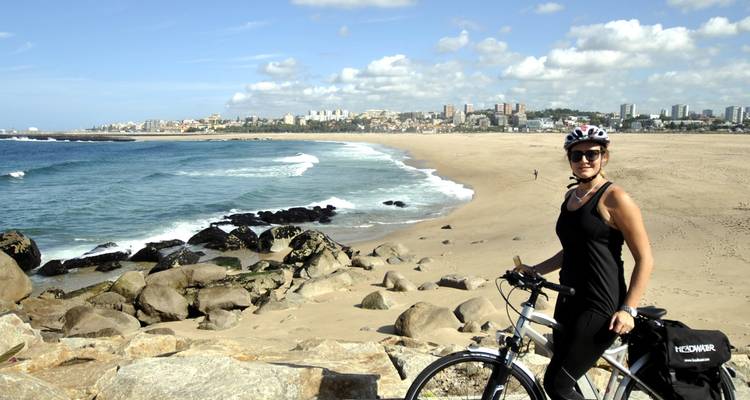 Ciclista posando con una bicicleta en una amplia playa de arena con olas del océano y el horizonte de Oporto al fondo.
