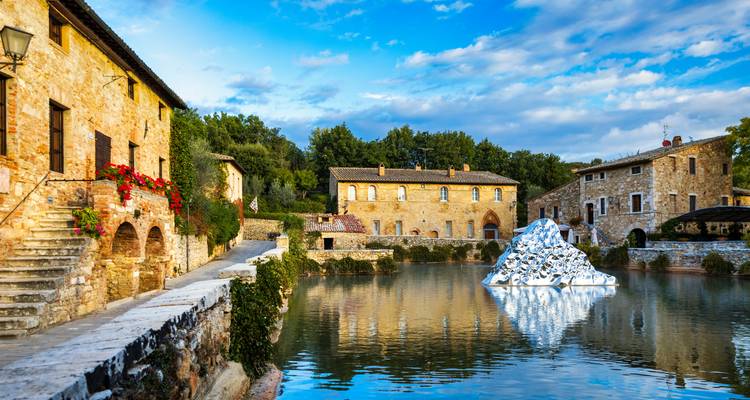 Bâtiments en pierre et piscine thermale de Bagno Vignoni se reflétant sous des ciels bleus.