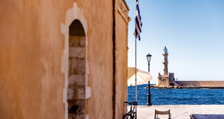 Vue du phare de La Canée à travers un port d'un bleu éclatant encadré par un ancien bâtiment en pierre et un drapeau grec.