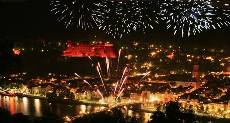 Vue nocturne d'une ville riveraine avec des feux d'artifice qui explosent au-dessus d'un château sur une colline illuminée.