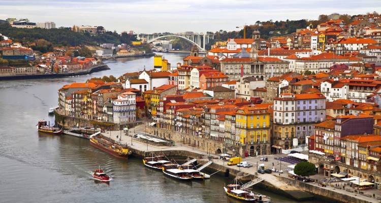 Panoramic view of a city with colorful buildings by a river and a bridge in the background.