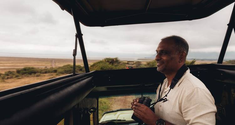 Ein Mann steht in einem offenen Safari-Fahrzeug und blickt über grasbewachsene Ebenen unter bewölktem Himmel.