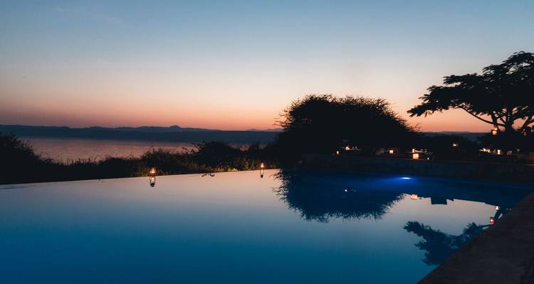 Ein ruhiger Infinity-Pool spiegelt die orangen und rosa Farbtöne des Sonnenuntergangs über einem See wider, mit kerzenbeschienenen Tischen am Rand.