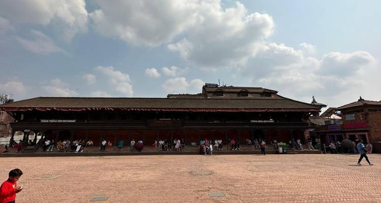 Wide plaza with people sitting and walking, bordered by traditional Nepalese architecture under a partly cloudy sky.