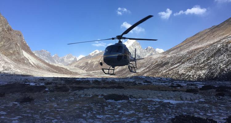 Helicopter hovering above snowy terrain with mountain peaks in the background.
