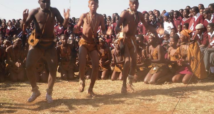 Traditionele dansers in leren outfit voeren een energieke routine uit voor een groot zittend publiek op een buitenveld.