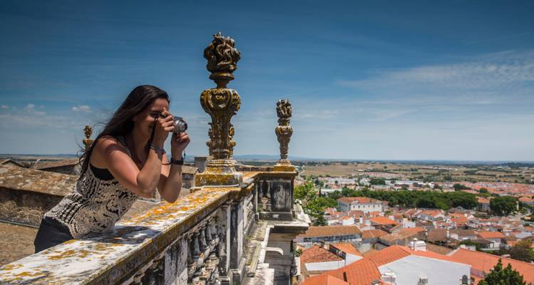 Mujer tomando una foto desde una balaustrada ornamentada que da vista a tejados de tejas rojas y el campo