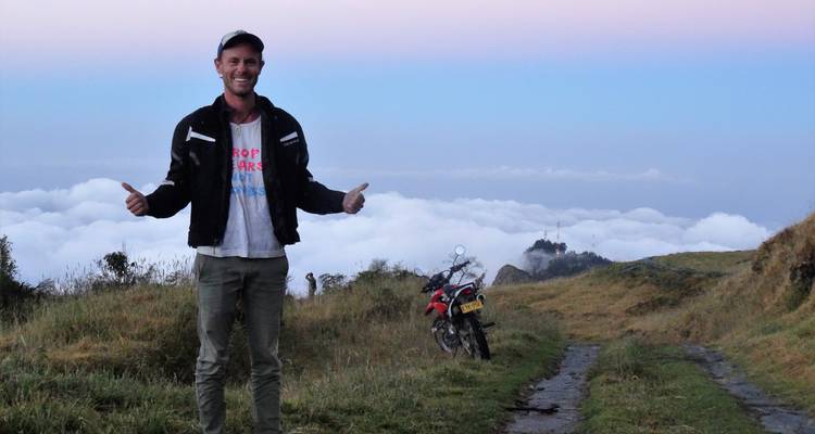 Man standing next to a motorbike overlooking clouds.