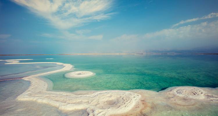 Buitenaardse zoutformaties omzomen het glasheldere turquoise water van de Dode Zee onder een mistige hemel.