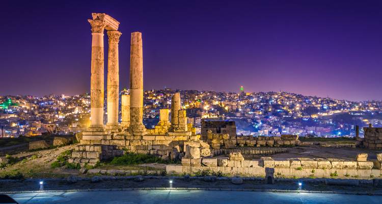 Columnas romanas iluminadas del Templo de Hércules contemplan el horizonte nocturno centelleante de Amán bajo un cielo púrpura profundo.
