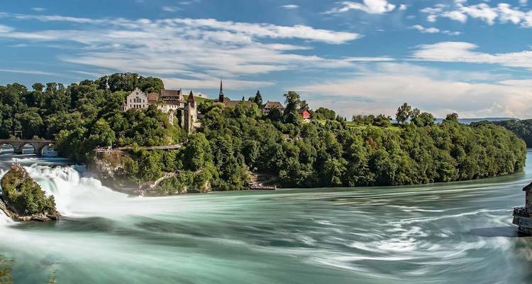 The powerful Rhine Falls cascades beside a forested hill crowned by a castle under a blue sky.